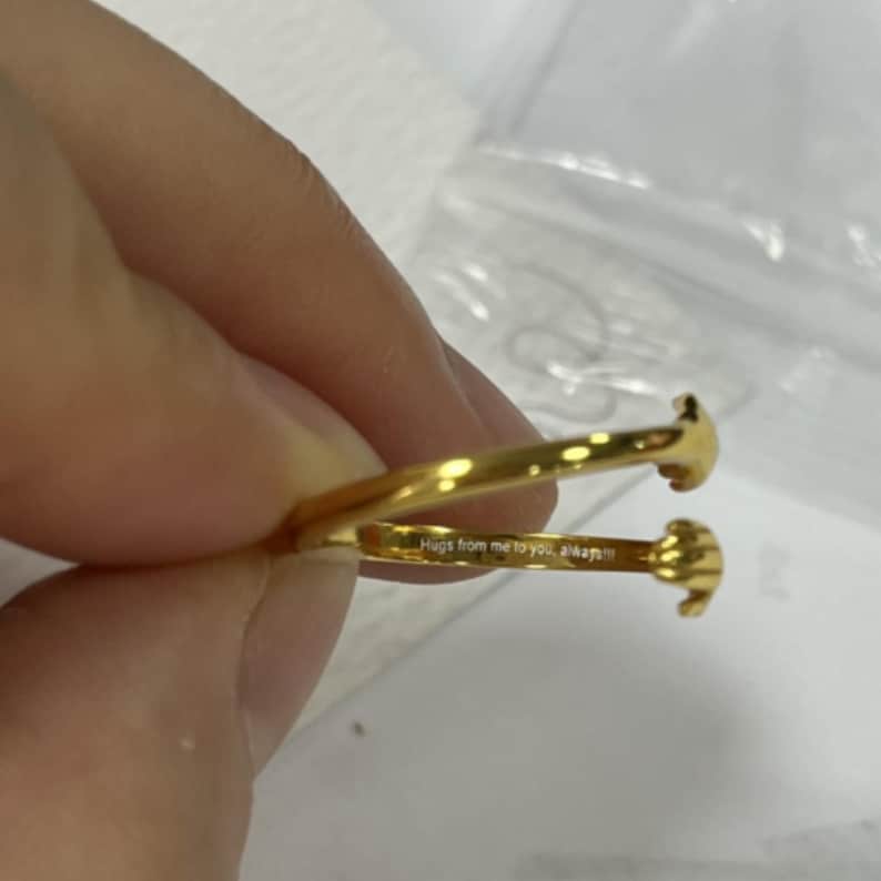 Puede incluir: Un anillo dorado con un dise&ntilde;o &uacute;nico. El anillo presenta dos manos estilizadas que parecen abrazarse. La inscripci&oacute;n "Hugs from me to you, always!!!" es visible en la banda del anillo.