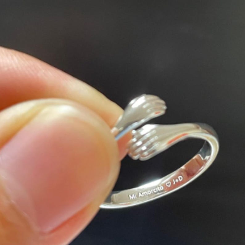 Puede incluir: Un anillo plateado con un dise&ntilde;o de dos manos abraz&aacute;ndose. El anillo est&aacute; sostenido por los dedos de una persona. El texto "Mi Amorcito J+D" est&aacute; grabado en el anillo.