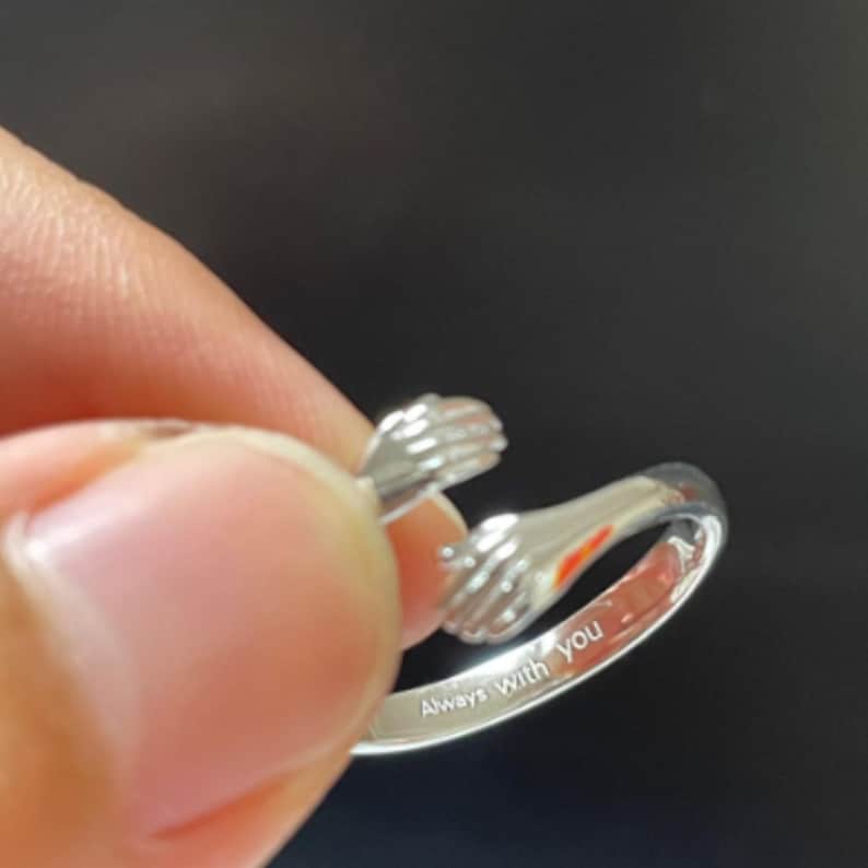 Puede incluir: Un anillo plateado con dos manos abraz&aacute;ndose. El anillo est&aacute; grabado con las palabras "Always With You". El anillo se muestra sobre un fondo oscuro.