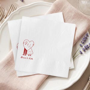 May include: Two white square napkins with a red heart design. The heart features the initials "G & L" and a stick figure couple. The words "Grace & Luke" are printed below the heart. The napkins are on a white plate with a pink napkin.