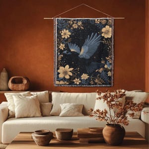 May include: A wall hanging depicting a dark blue bird in flight, framed by yellow and white flowers and leaves against a starry night. The fringed tapestry is suspended from a wooden rod, bringing a natural artistic element to the space.