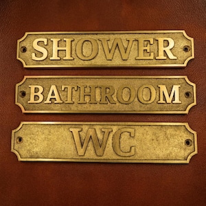 May include: Three brass bathroom signs with the words "SHOWER", "BATHROOM", and "WC" embossed on them. The signs are rectangular with decorative edges and pre-drilled holes for mounting. The background is a dark brown leather.