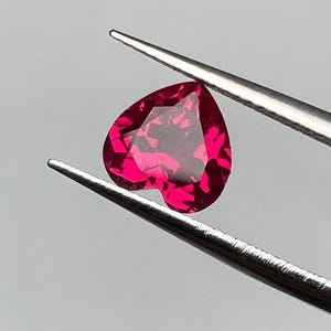 Puede incluir: Una gema facetada en forma de corazón, de un tono rojo vibrante, es sostenida por unas pinzas plateadas. El corte de la gema revela múltiples facetas, creando un efecto brillante. El fondo es de un gris suave y neutro.