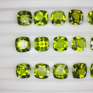 May include: A collection of cushion-cut, olive-green gemstones. The gems are arranged in rows, each displaying facets that catch the light. The stones have a vibrant, yellowish-green hue, suggesting peridot or a similar gemstone. The overall presentation is clean and organized.