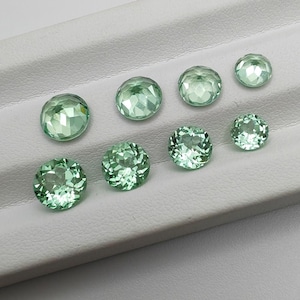 May include: A collection of eight faceted, round, light green gemstones of varying sizes. The gems are arranged on a white, textured surface, showcasing their clarity and cut. The stones have a sparkling, translucent appearance.
