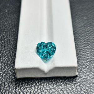 Peut inclure: Une pierre précieuse taillée en forme de cœur, de couleur turquoise vif. La gemme est présentée sur une surface blanche, sur un fond noir texturé. La taille de la pierre crée un effet scintillant, mettant en valeur son éclat.