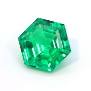 Puede incluir: Una gema verde esmeralda facetada, de forma geométrica. La gema tiene una superficie brillante y reflectante, sobre un fondo blanco. Las facetas de la gema crean un juego de luces y sombras, resaltando su claridad y color.