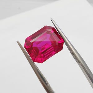 Könnte beinhalten: Ein facettierter, rechteckig geschliffener Rubin-Edelstein, der von einer Pinzette gehalten wird. Der Rubin hat eine leuchtende, tiefrosa Farbe mit mehreren Facetten, die das Licht reflektieren. Die Pinzette ist silberfarben, der Hintergrund ist weiß.