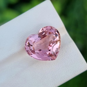 May include: A heart-shaped, faceted gemstone in a soft pink hue. The gem is held on a white surface, with a blurred green background. The gemstone's facets catch the light, creating a sparkling effect. This could be a pendant or a loose gemstone.