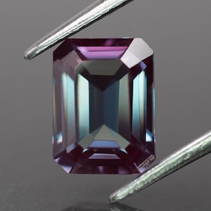 Può includere: Una gemma taglio rettangolare con effetto cangiante, tenuta da pinzette. La gemma passa dal viola al verde acqua e blu, con una superficie sfaccettata che riflette la luce. La gemma è al centro di un sfondo neutro.