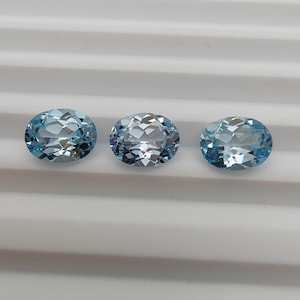 May include: Three oval-cut, light blue gemstones. Each gem has a faceted surface, reflecting light. The gems are arranged in a row against a white, ridged background. These gemstones are suitable for jewelry making.