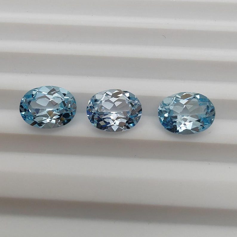 May include: Three oval-cut, light blue gemstones. Each gem has a faceted surface, reflecting light. The gems are arranged in a row against a white, ridged background. These gemstones are suitable for jewelry making.