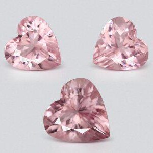May include: Three heart-shaped, faceted gemstones in a soft pink hue. The gems are arranged on a light background, showcasing their intricate cuts and reflective surfaces. The gems are likely for jewelry making or collecting.