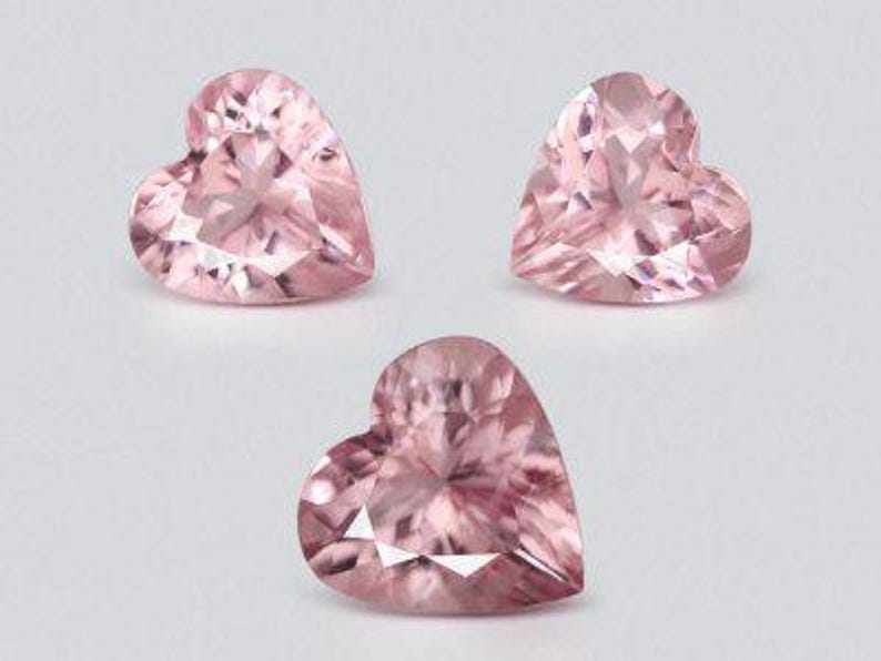 May include: Three heart-shaped, faceted gemstones in a soft pink hue. The gems are arranged on a light background, showcasing their intricate cuts and reflective surfaces. The gems are likely for jewelry making or collecting.
