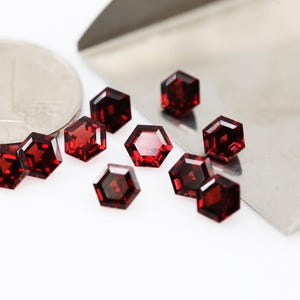 Può includere: Primo piano di diverse pietre preziose esagonali sfaccettate di colore rosso intenso. Le gemme sono sparse su una superficie bianca, con uno strumento d'argento e una moneta sullo sfondo. Le gemme hanno una superficie lucida e riflettente.