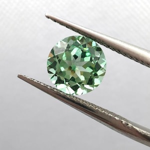 Könnte beinhalten: Ein runder, facettierter Edelstein in Grüntönen wird von einer silbernen Pinzette gehalten. Der Stein hat einen Brillantschliff, der das Licht reflektiert und seine innere Struktur zeigt. Der Hintergrund ist ein weiches, neutrales Weiß.