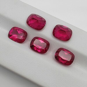 Könnte beinhalten: Fünf facettierte, kissenförmige, rubinrote Edelsteine liegen auf einer weißen Oberfläche. Die Edelsteine haben eine hohe Klarheit und Brillanz und reflektieren das Licht. Die Steine sind tiefrot, rechteckig und haben abgerundete Ecken.