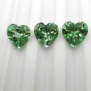 Op de afbeelding: Drie hartvormige groene edelstenen. Elke edelsteen heeft meerdere facetten, waardoor een sprankelend effect ontstaat. De edelstenen zijn in een rij gerangschikt tegen een witte achtergrond. De edelstenen hebben een levendige, lichtgroene kleur.