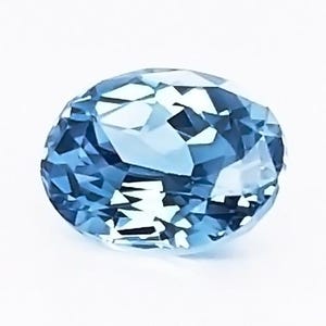 May include: An oval-shaped, faceted gemstone in shades of light blue. The gem has a brilliant cut, reflecting light and creating a sparkling effect. The stone is set against a white background, highlighting its color and clarity.