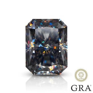 Puede incluir: Una gema de corte rectangular, gris oscuro, con múltiples facetas, que refleja un espectro de colores. La gema está sobre un fondo blanco, con las letras "GRA" y un logotipo circular visibles.