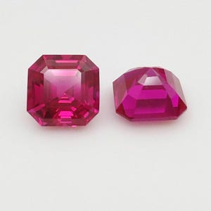 Könnte beinhalten: Zwei facettierte Edelsteine im Radiant-Schliff mit tiefem Magenta-Farbton. Einer ist quadratisch, der andere im modifizierten Kissenschliff. Die Edelsteine sind vor weißem Hintergrund platziert, was ihre Brillanz und Klarheit hervorhebt. Ideal für Schmuck.