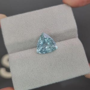 May include: A light blue, triangular gemstone rests on a gray, textured surface. The gemstone has a faceted surface, catching the light. The stone is held between two fingers, suggesting a jewelry or gemstone display.