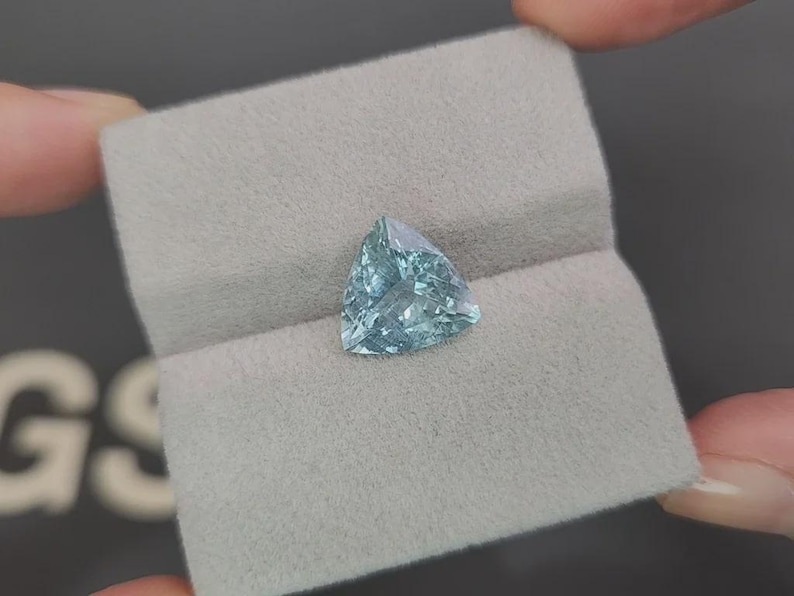 May include: A light blue, triangular gemstone rests on a gray, textured surface. The gemstone has a faceted surface, catching the light. The stone is held between two fingers, suggesting a jewelry or gemstone display.