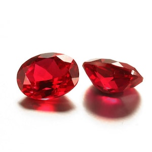 Könnte beinhalten: Zwei facettierte, oval geformte, leuchtend rote Edelsteine. Einer ist aufrecht positioniert, während der andere auf der Seite liegt und den Schliff zeigt. Die Edelsteine haben einen hohen Glanz und sind vor weißem Hintergrund platziert.