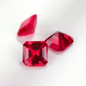 Könnte beinhalten: Drei facettierte, leuchtend rote Edelsteine ​​werden vor einem weißen Hintergrund präsentiert. Der größte Edelstein ist quadratisch mit abgeschrägten Ecken, während die anderen unterschiedliche Formen haben. Die Edelsteine ​​sind hochreflektierend und zeigen ihre innere Struktur.