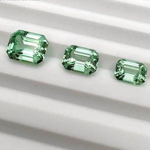 May include: Three green emerald-cut gemstones of different sizes. The gems have a faceted surface that reflects light. The stones are arranged in a row on a white surface. The gems are in a row.