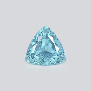 May include: A faceted, triangular-cut gemstone with a light blue hue. The gem has a symmetrical shape with multiple facets that reflect light, creating a sparkling effect. The gemstone is set against a plain, light gray background.