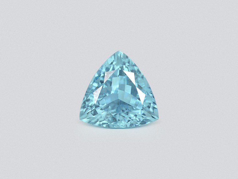 May include: A faceted, triangular-cut gemstone with a light blue hue. The gem has a symmetrical shape with multiple facets that reflect light, creating a sparkling effect. The gemstone is set against a plain, light gray background.