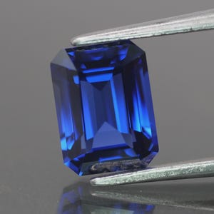 Op de afbeelding: Een diepblauwe edelsteen met rechthoekige slijpvorm wordt vastgehouden door een zilveren pincet. De edelsteen heeft een glanzend, reflecterend oppervlak dat de interne structuur en facetten laat zien. De achtergrond is neutraal grijs.