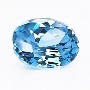May include: A faceted, oval-shaped gemstone with a light blue hue. The gem has multiple facets that reflect light, creating a sparkling effect. The gemstone is set against a white background.