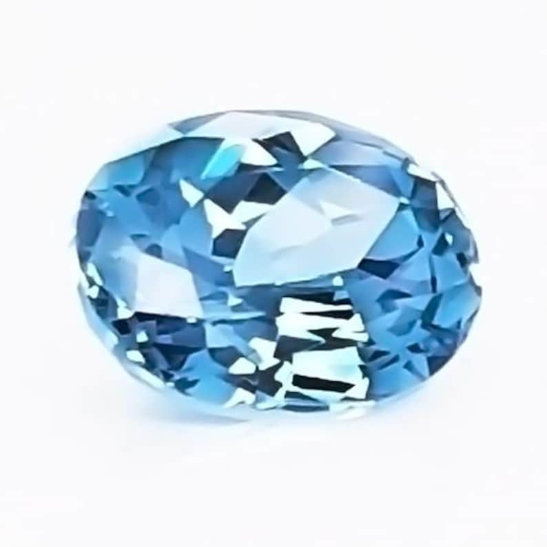 May include: A faceted, oval-shaped gemstone with a light blue hue. The gem has multiple facets that reflect light, creating a sparkling effect. The gemstone is set against a white background.