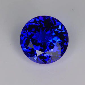 Könnte beinhalten: Ein runder, facettierter Edelstein in tiefblauer Farbe. Die Oberfläche des Edelsteins reflektiert das Licht und erzeugt einen funkelnden Effekt. Der Edelstein ist vor einem neutralen Hintergrund platziert, um seine Farbe und Brillanz hervorzuheben. Geeignet für Schmuck.