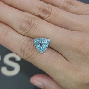 May include: A triangular, light blue gemstone with a faceted surface. The gem is held in a hand, showcasing its clarity and color. The stone's shape is a prominent feature, reflecting light and creating a sparkling effect. The gem is likely a piece of jewelry.