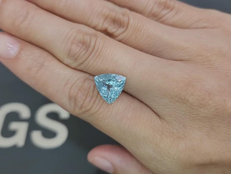 May include: A triangular, light blue gemstone with a faceted surface. The gem is held in a hand, showcasing its clarity and color. The stone's shape is a prominent feature, reflecting light and creating a sparkling effect. The gem is likely a piece of jewelry.