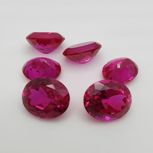 Könnte beinhalten: Eine Ansammlung von facettierten, ovalen und runden Edelsteinen in verschiedenen Größen. Die Edelsteine haben eine tiefe, lebendige magentafarbene Farbe und sind auf einer weißen Oberfläche angeordnet.