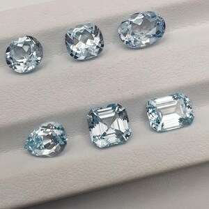 May include: A collection of six aquamarine gemstones in various shapes, including oval, cushion, and emerald cuts. The gems are a light blue color and are displayed on a white surface.