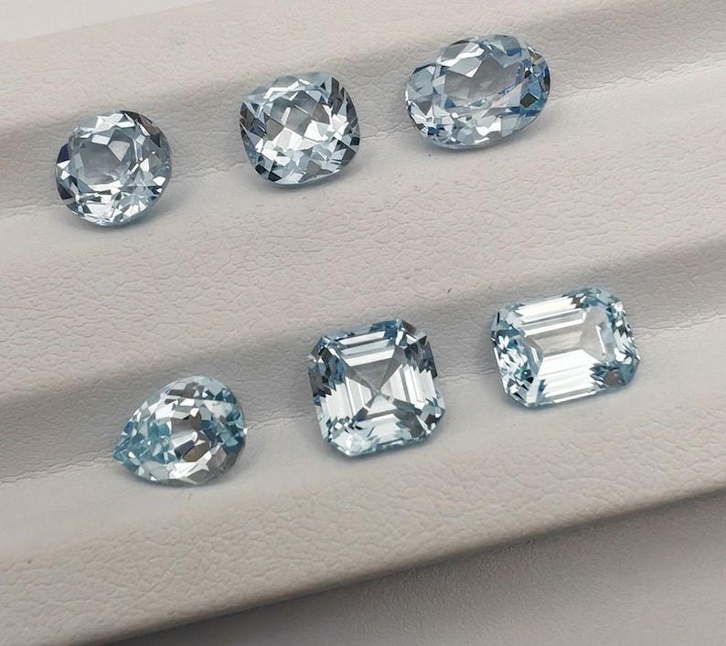 May include: A collection of six aquamarine gemstones in various shapes, including oval, cushion, and emerald cuts. The gems are a light blue color and are displayed on a white surface.