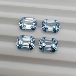 Peut inclure: Quatre pierres précieuses de couleur bleu clair, de taille émeraude. Chaque gemme a une forme rectangulaire avec des facettes taillées, reflétant la lumière. Les gemmes sont disposées sur une surface blanche. Ces pierres sont idéales pour la joaillerie.