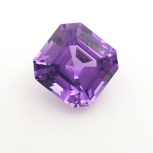 Könnte beinhalten: Ein facettierter, quadratisch geschliffener Amethyst-Edelstein. Der Edelstein hat eine tiefviolette Farbe mit mehreren Facetten, die das Licht reflektieren. Der Edelstein ist vor einem weißen Hintergrund platziert.