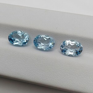 May include: Three oval-cut, light blue gemstones are displayed. The gems have a faceted surface, reflecting light. The stones are arranged in a row against a white background, showcasing their clarity and color. These gemstones are suitable for jewelry making.