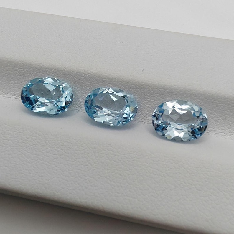 May include: Three oval-cut, light blue gemstones are displayed. The gems have a faceted surface, reflecting light. The stones are arranged in a row against a white background, showcasing their clarity and color. These gemstones are suitable for jewelry making.