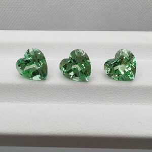 Op de afbeelding: Drie hartvormige, gefacetteerde groene edelstenen. De edelstenen worden gepresenteerd op een wit oppervlak, waarbij hun ingewikkelde slijpsels en reflecterende eigenschappen worden benadrukt. De edelstenen zijn waarschijnlijk voor het maken van sieraden.