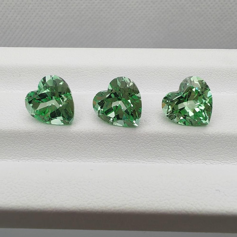 Op de afbeelding: Drie hartvormige, gefacetteerde groene edelstenen. De edelstenen worden gepresenteerd op een wit oppervlak, waarbij hun ingewikkelde slijpsels en reflecterende eigenschappen worden benadrukt. De edelstenen zijn waarschijnlijk voor het maken van sieraden.