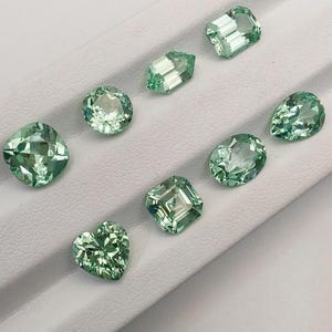 May include: A collection of eight faceted gemstones in various shapes, including round, oval, pear, and heart. The gems are a light green color and are displayed on a white surface. The gemstones are cut to reflect light.