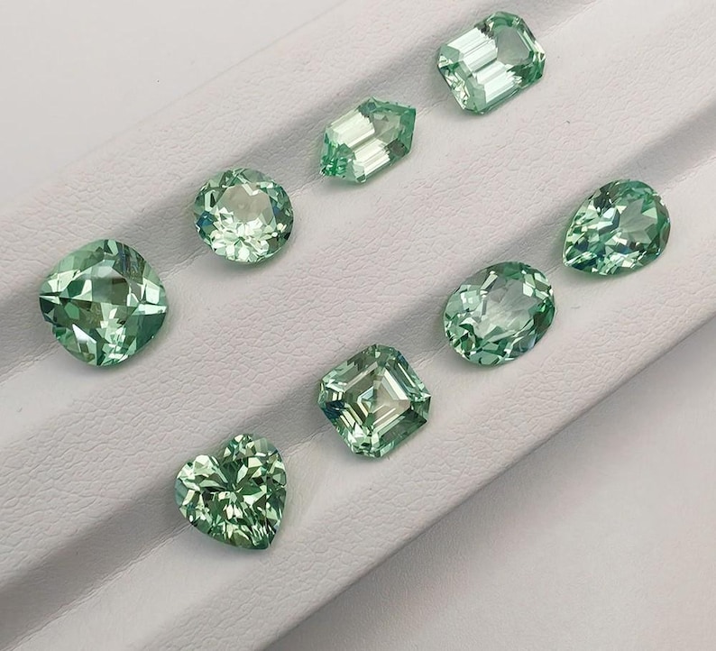 May include: A collection of eight faceted gemstones in various shapes, including round, oval, pear, and heart. The gems are a light green color and are displayed on a white surface. The gemstones are cut to reflect light.
