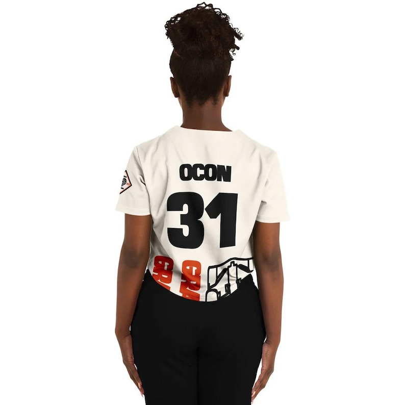 May include: Cream-colored t-shirt with the name "OCON" and the number "31" in black. The bottom of the shirt features a graphic design with red and black elements. The person is wearing black pants.
